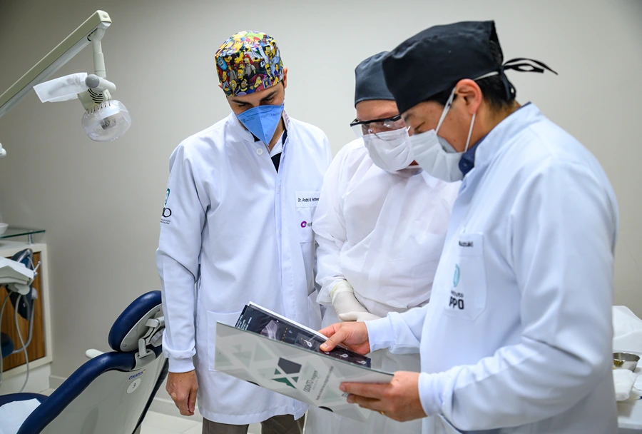 Três profissionais da odontologia, em trajes de proteção, analisando um material impresso durante uma sessão prática de implantodontia.