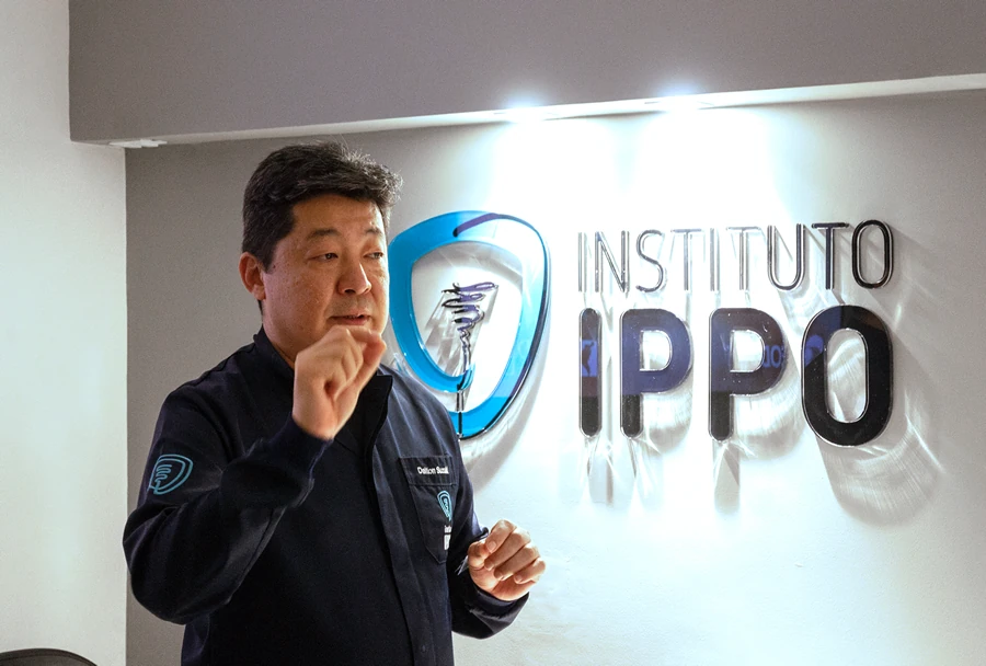 Professor Dalton Suzuki do Instituto IPPO, em frente ao logo do instituto, explicando um conceito para os alunos durante uma apresentação.