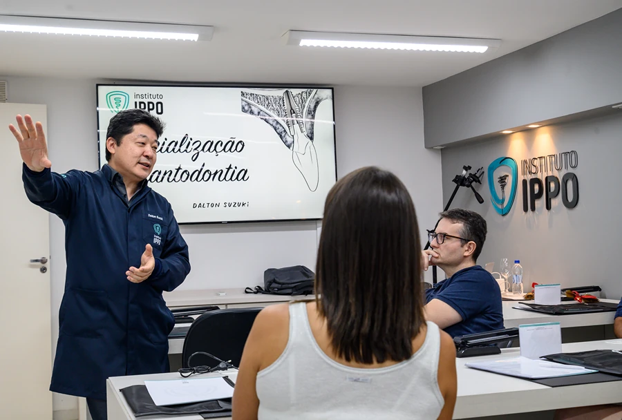 Instrutor explicando o conteúdo de especialização em Implantodontia aos alunos, com slides no fundo mostrando uma imagem de anatomia dental.