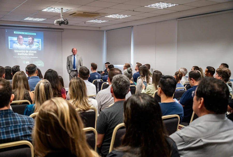 Homem do IPPO em terno azul falando em palestra, público em cadeiras, slide com fotos e texto sobre encontro de alunos.