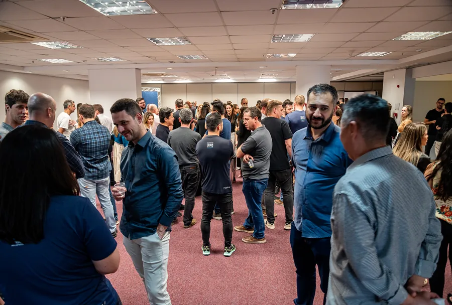 Grande grupo do IPPO conversando em networking, sala com carpete vermelho, teto com luminárias, alguns em camisetas azuis com logo.