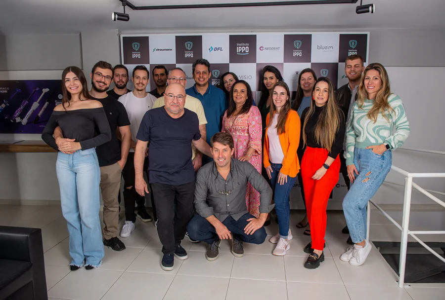 Grupo de pessoas de várias idades posando juntas em frente ao painel de patrocinadores do Instituto IPPO, algumas vestindo roupas casuais, todos sorrindo para a câmera.