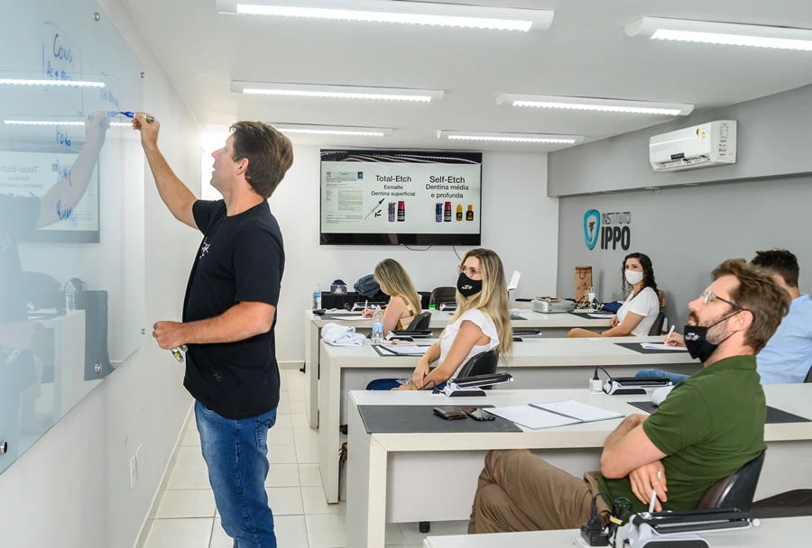 Instrutor escrevendo na lousa enquanto alunos mascarados prestam atenção, com informações projetadas na tela sobre "Total Etch" e "Self-Etch".