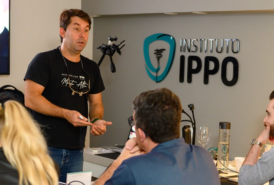 Professor usando camiseta preta gesticulando enquanto fala para alunos do Instituto IPPO, com o logotipo do instituto ao fundo.