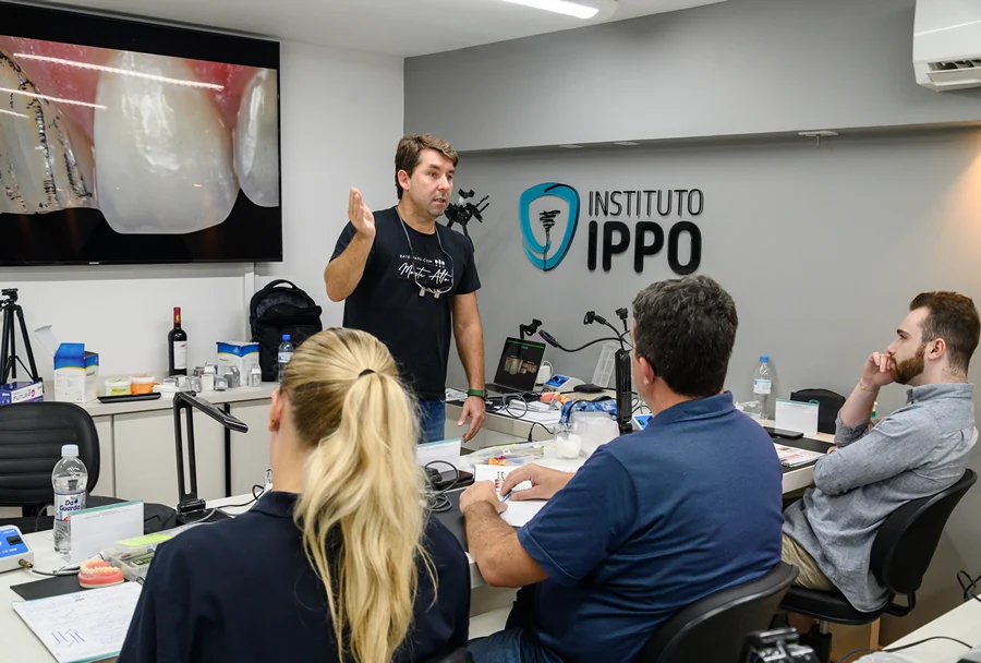 Professor em camiseta preta apresentando e explicando um procedimento odontológico com imagem ampliada de um dente na tela.