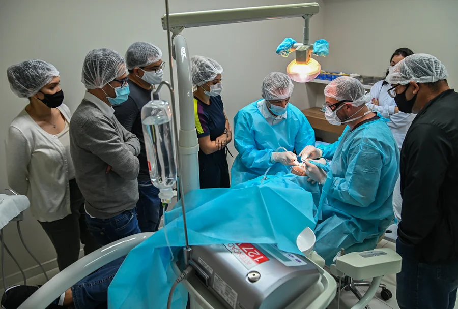 Aula prática em um ambiente clínico, onde alunos e o professor realizam uma cirurgia guiada sob um foco cirúrgico, todos equipados com aventais e toucas cirúrgicas.