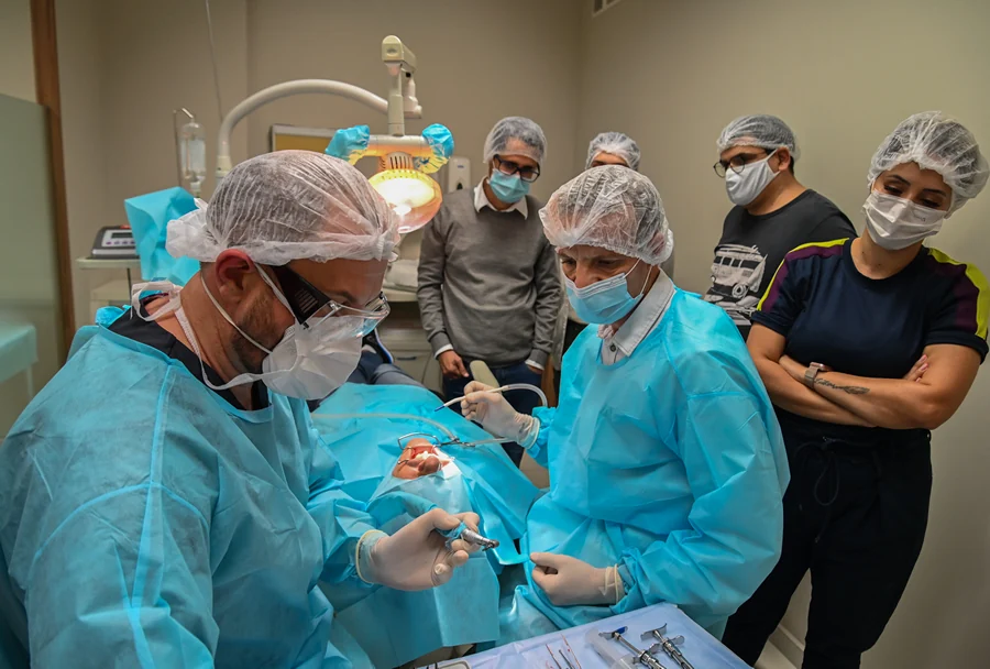 Alunos observando atentamente uma cirurgia guiada sendo realizada pelo professor, com um ambiente cirúrgico completo e todos os envolvidos equipados adequadamente.