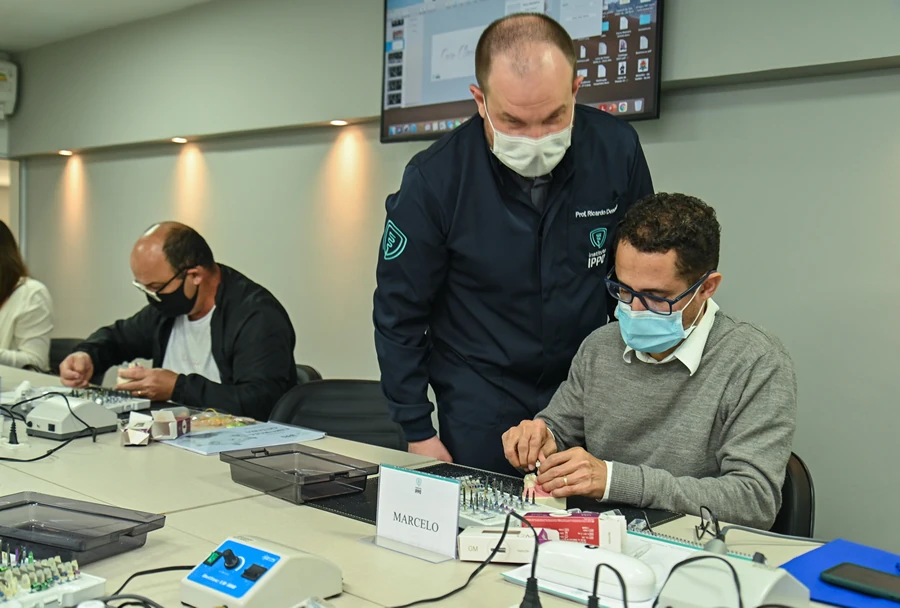 Instrutor auxiliando um participante na realização de uma técnica prática de cirurgia guiada, ambos utilizando máscaras de proteção.