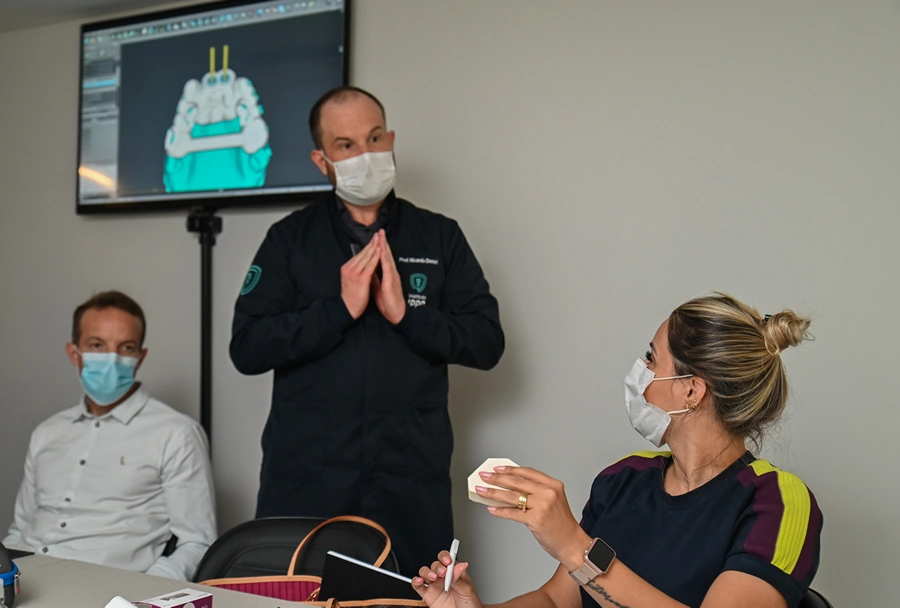 Participantes prestando atenção em uma explicação enquanto observam o instrutor apresentar uma modelagem 3D de um procedimento odontológico no telão.