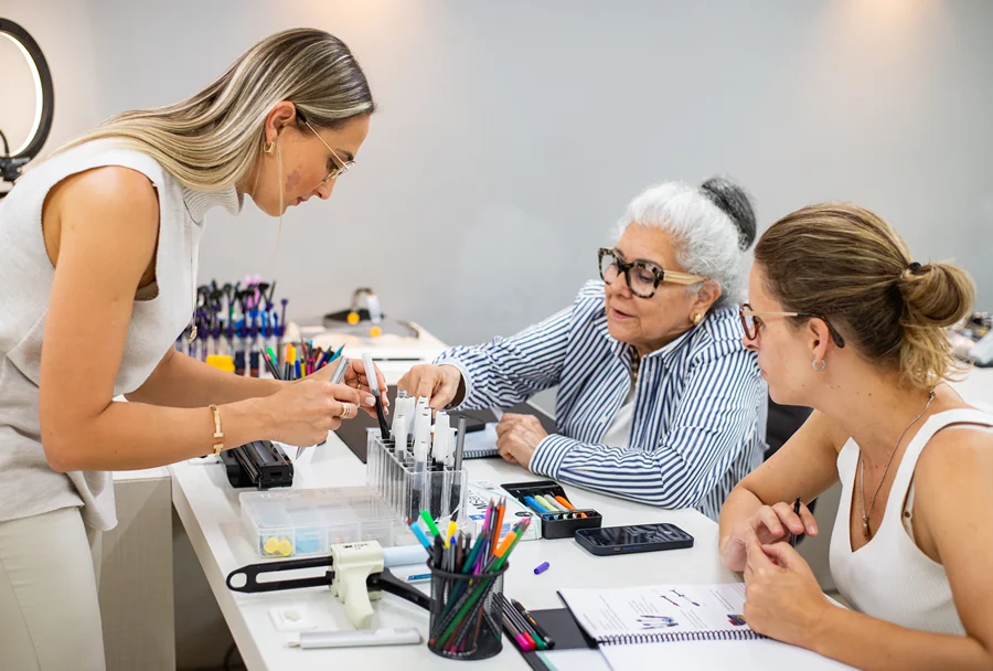 Instrutora auxiliando duas participantes em um exercício prático com materiais odontológicos e marcadores coloridos na mesa.